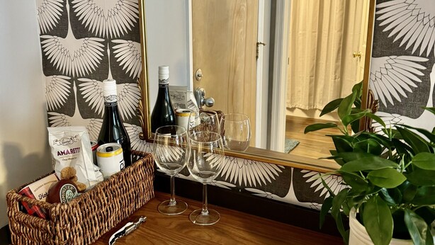 Ein Zimmer mit einem hölzernen Schminktisch, einem Spiegel und einer Wand mit einem Blattmuster. Auf dem Schminktisch stehen Weingläser, Flaschen und ein Korb.