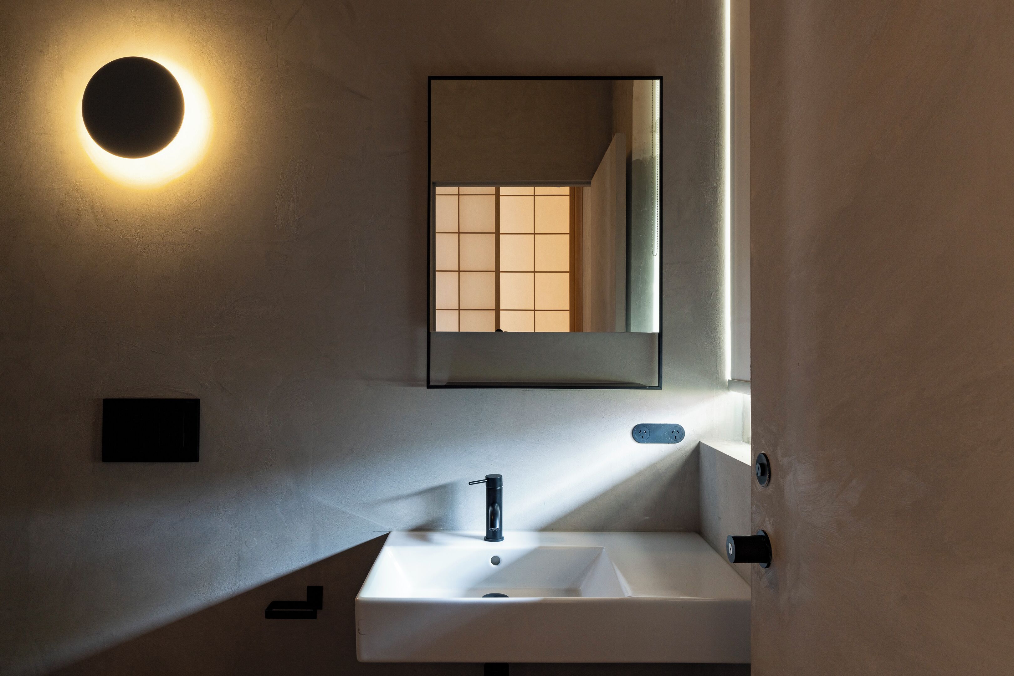A modern bathroom with a white sink, a round mirror, and a wall-mounted light fixture.