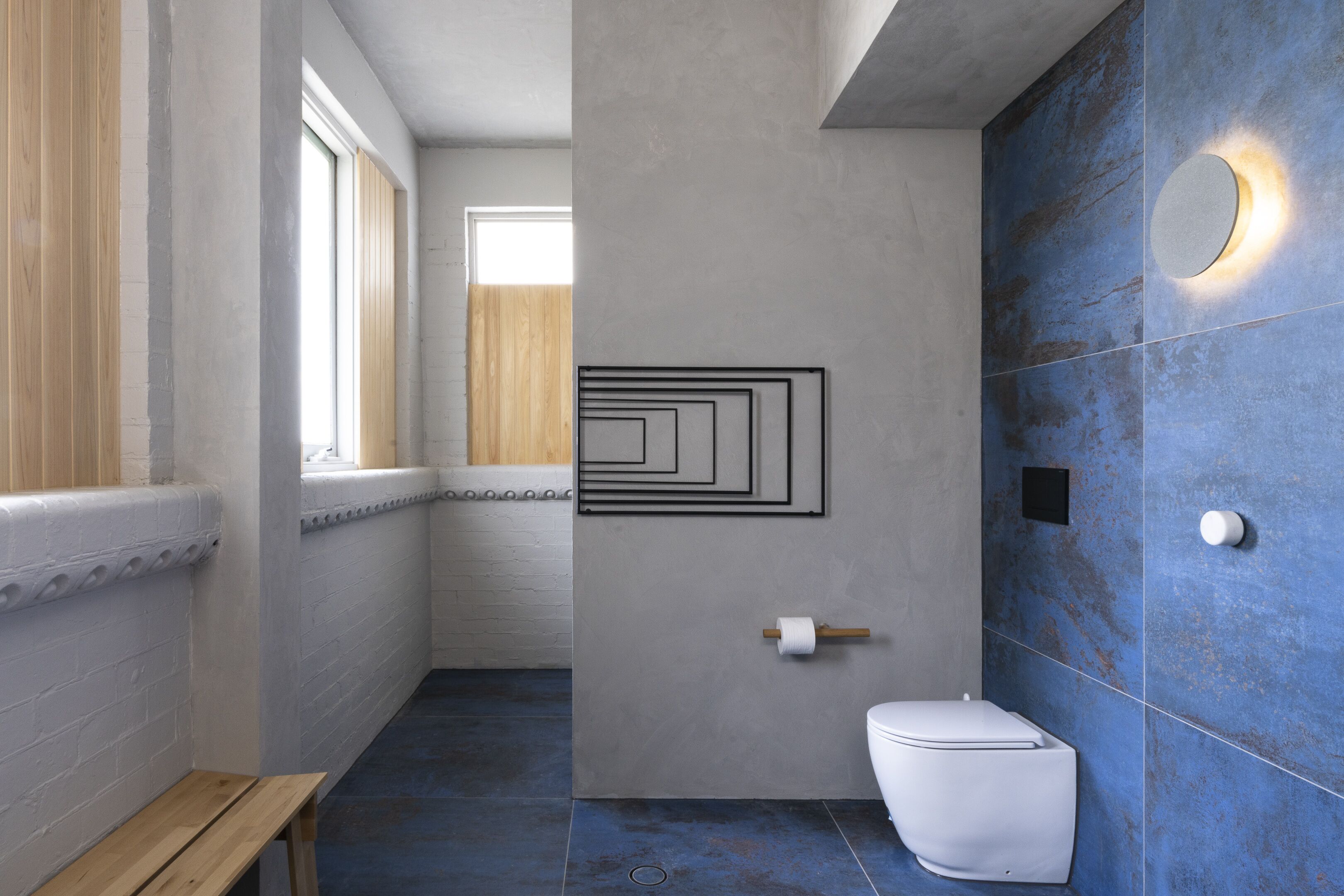 Un baño con inodoro blanco, un banco de madera, pared con azulejos azules y una ventana.