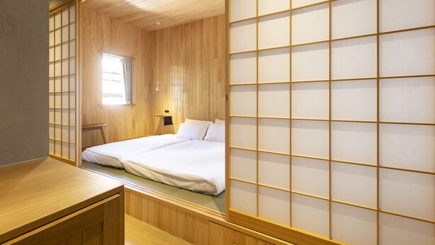 Une pièce de style japonais traditionnel, avec un plancher en bois, un lit recouvert de draps blancs, un paravent coulissant de type shoji et une petite fenêtre avec des rideaux.