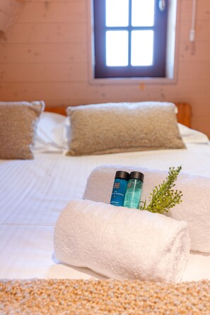 Una cama bien hecha con sábanas blancas, dos botellas de productos de baño y una ramita de verdor sobre una toalla beige con textura.
