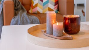 A cozy living room with a white table holding candles and a tray, a brown leather sofa with colorful pillows, and a modern kitchen in the background.