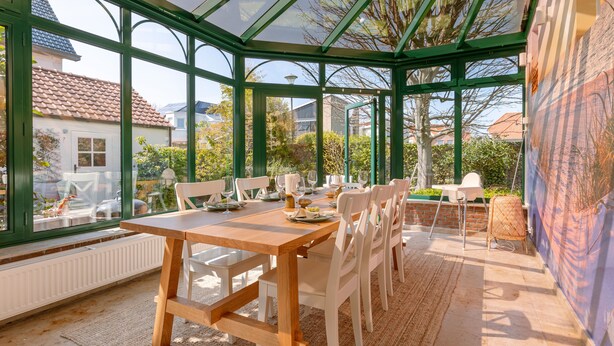 A conservatory with a glass roof, a wooden table set for dining, and a view of the surrounding garden and houses.