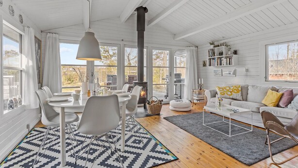Ett rymligt vardagsrum med högt tak, vita träväggar, en centralt placerad öppen spis och en matplats med moderna stolar och en mönstrad matta.