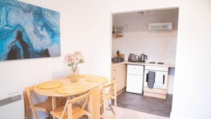 A compact kitchen with white cabinets, a small refrigerator, and a stove. A dining table with chairs is positioned in front of a large abstract painting. - Lovely 1 bed Apt - Buckinghamshire (High Wycombe)