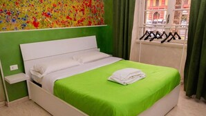 Una cama bien hecha con cubrecama verde y almohadas blancas, una mesita de noche blanca con un pequeño estante, y una obra de arte floral colorida encima de la cama.
