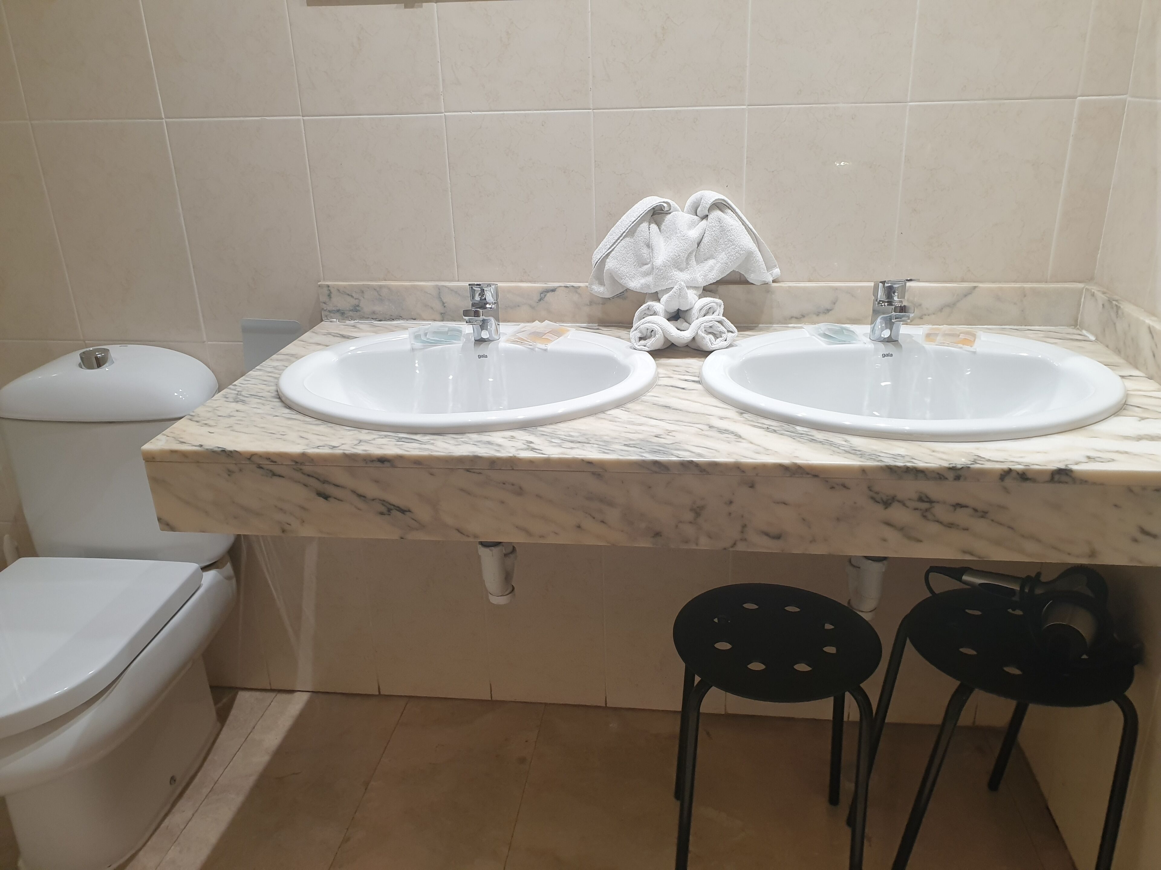 A bathroom with two sinks, a toilet, and two black chairs.