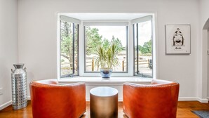 Ett rum med ett stort fönster, två läderbeklädda sittpuffar, en vas av metall och en inramad tavla på väggen.