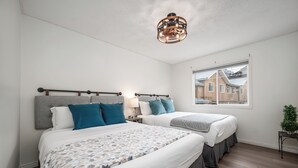 A bedroom with two beds, a window with a view of houses, a ceiling light fixture, and a small plant on a side table. - Luxurious Retreat/Hot Tub/Mountain View/4 Beds (Bighorn No. 8)