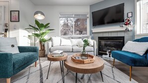 A modern living room with a grey sofa, teal armchair, and blue armchair. There is a fireplace, a large TV, and a potted plant. - Luxurious Retreat/Hot Tub/Mountain View/4 Beds (Bighorn No. 8)