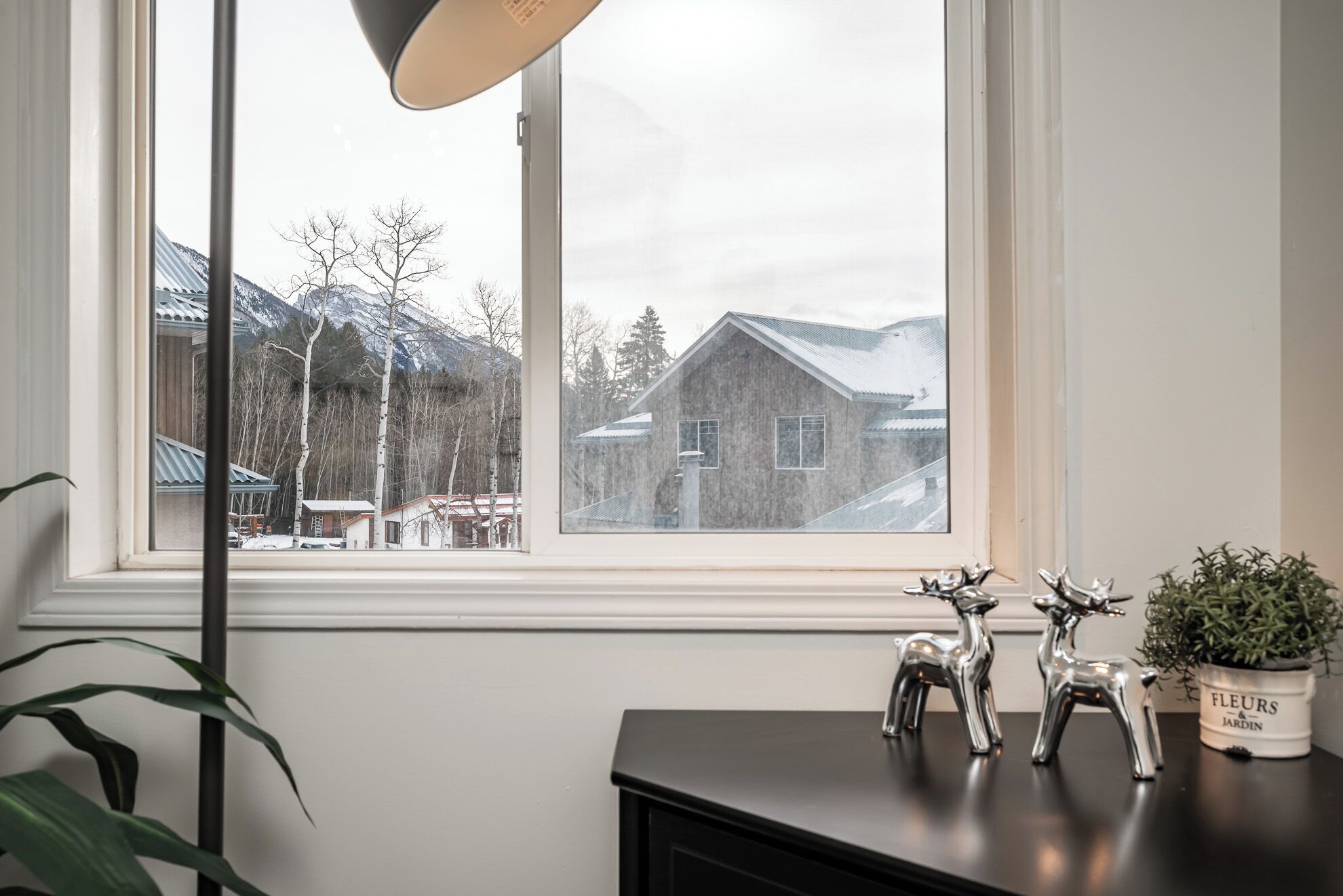 Habitación con vistas a una montaña nevada y una casa de madera. Se aprecia una ventana con marco blanco, una mesa negra con objetos decorativos y una planta en maceta.