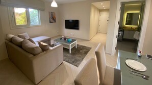 Un salon moderne avec un canapé, une table basse et un téléviseur. On y trouve également un coin repas avec une table dressée pour deux personnes. La pièce est dotée d’une grande fenêtre et d’une porte donnant sur une salle de bain.