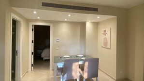 Un espace repas moderne avec une table en verre et deux chaises, une bouche d’aération au plafond et une porte donnant sur une chambre.