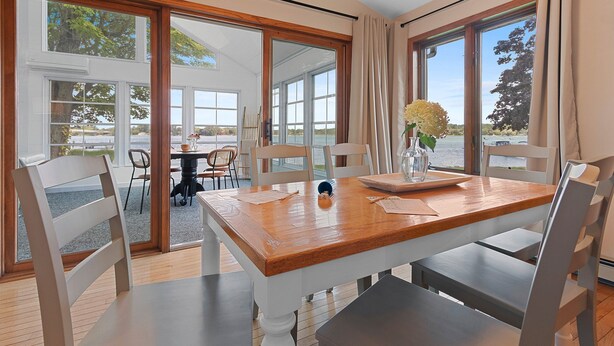 Un comedor con mesa de madera y sillas blancas, ventanas amplias con cortinas y vistas al exterior.