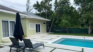A swimming pool with two lounge chairs and a covered umbrella, surrounded by a paved patio and a house with a green exterior. - House by the lake: 3BR/2.5BA, Gated with a Pool! (Nassau)