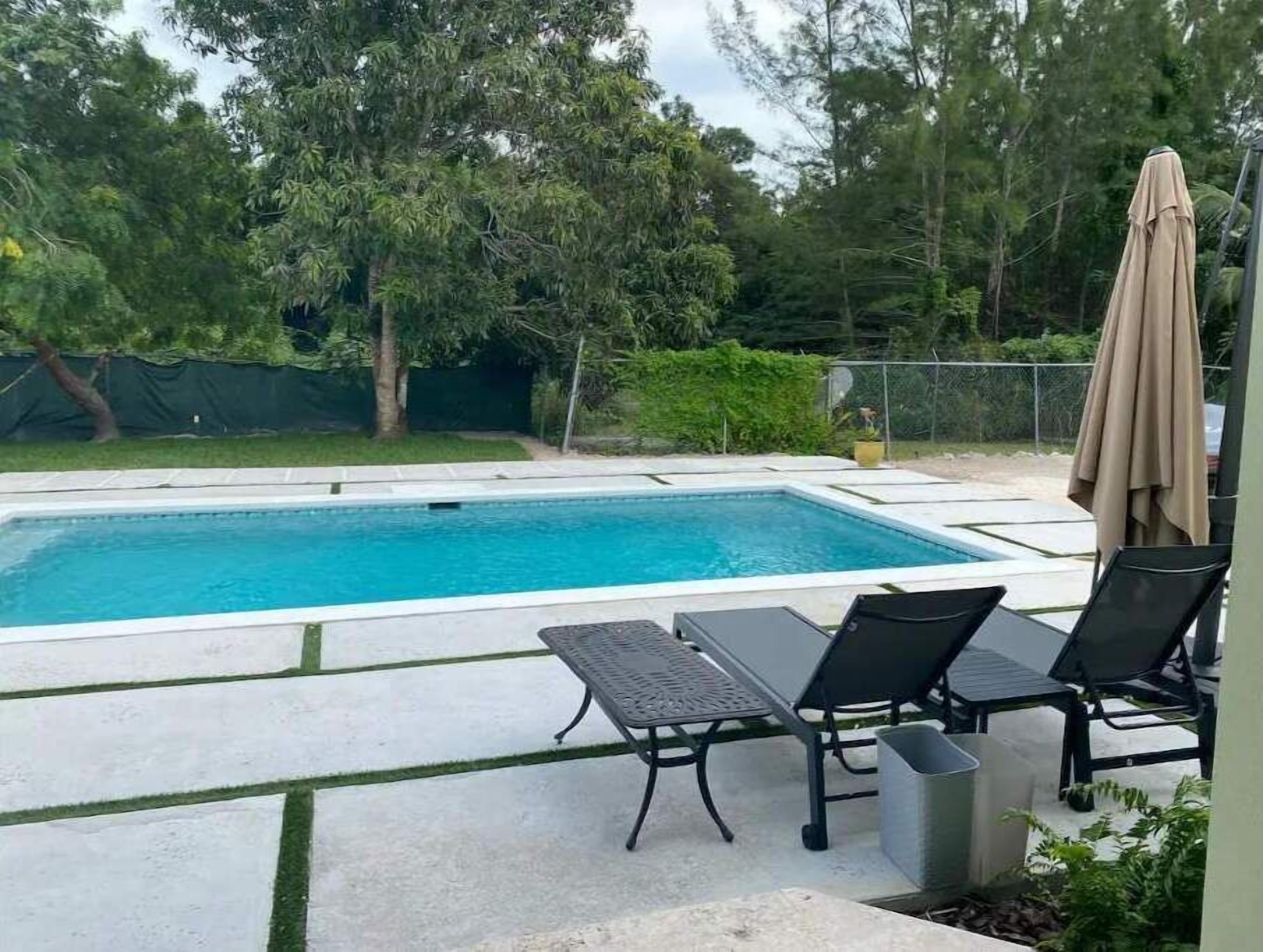 A rectangular swimming pool with a tiled deck, surrounded by greenery and trees. There are two lounge chairs and a table with a metal frame.