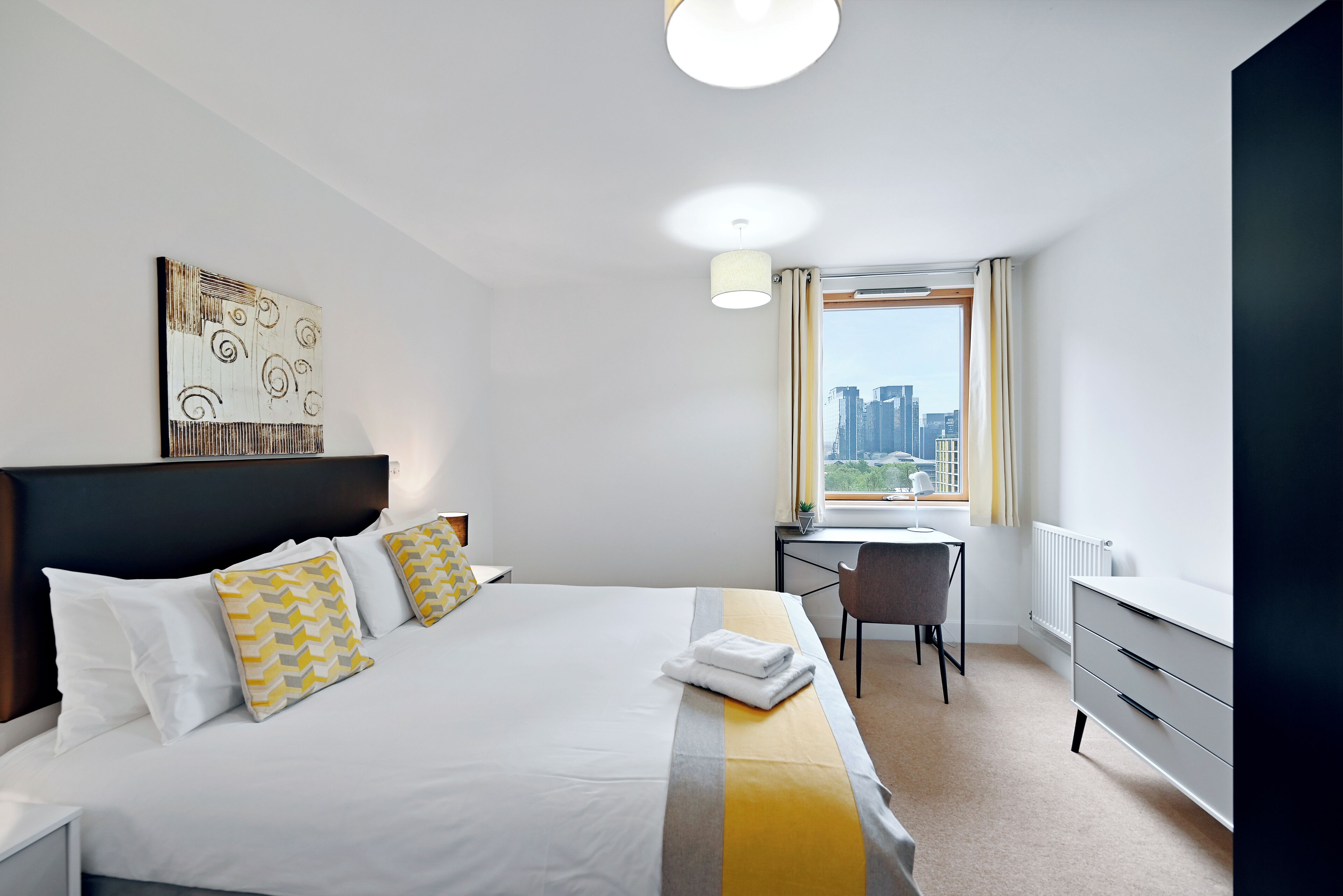 A hotel room with a bed, a desk, a chair, and a view of the city.