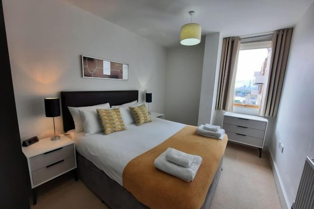 A hotel room with a bed, bedside tables, a lamp, a window with curtains, and a city view.