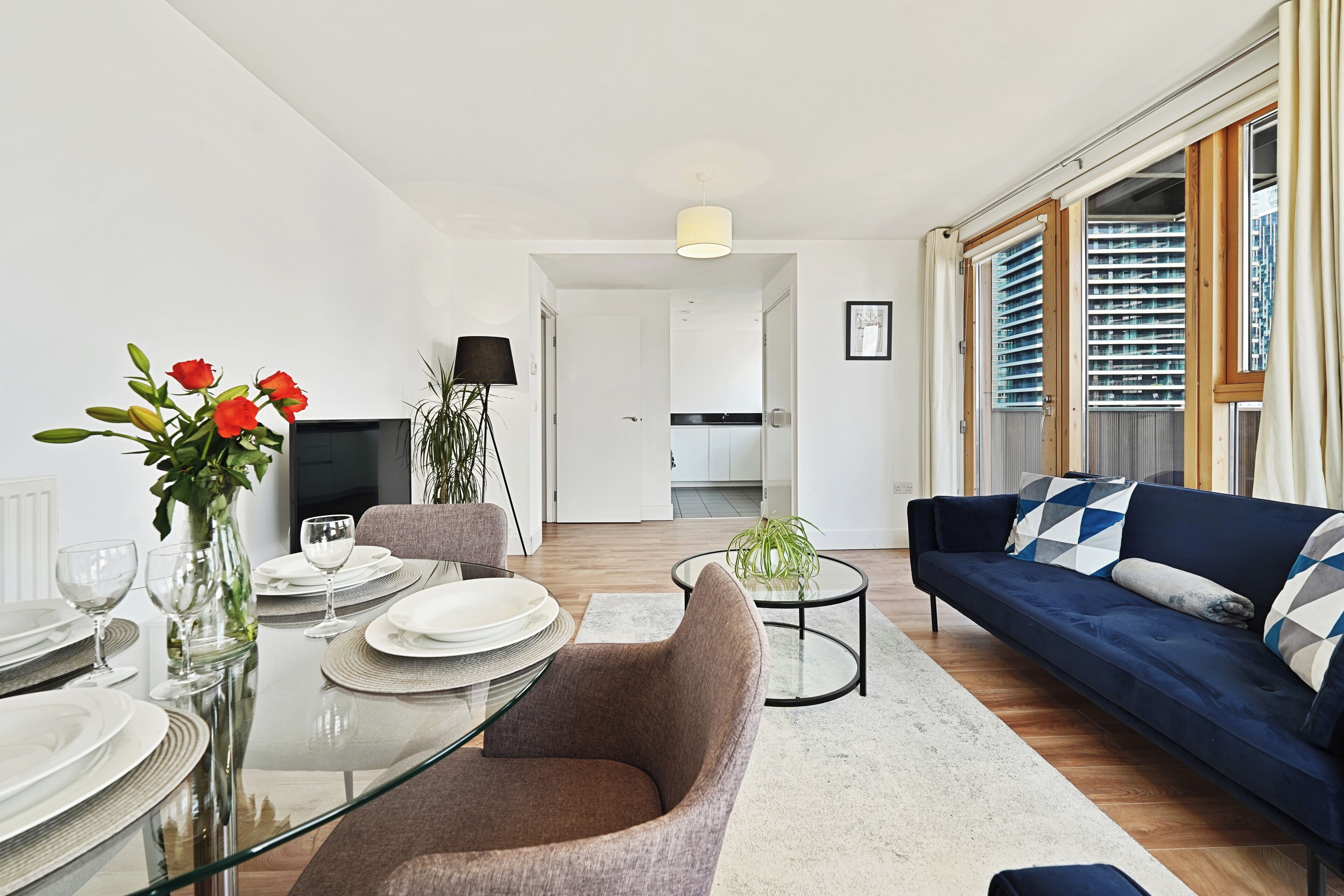 A modern living room with a glass dining table set for four, a blue sofa, and a view of the city through large windows.