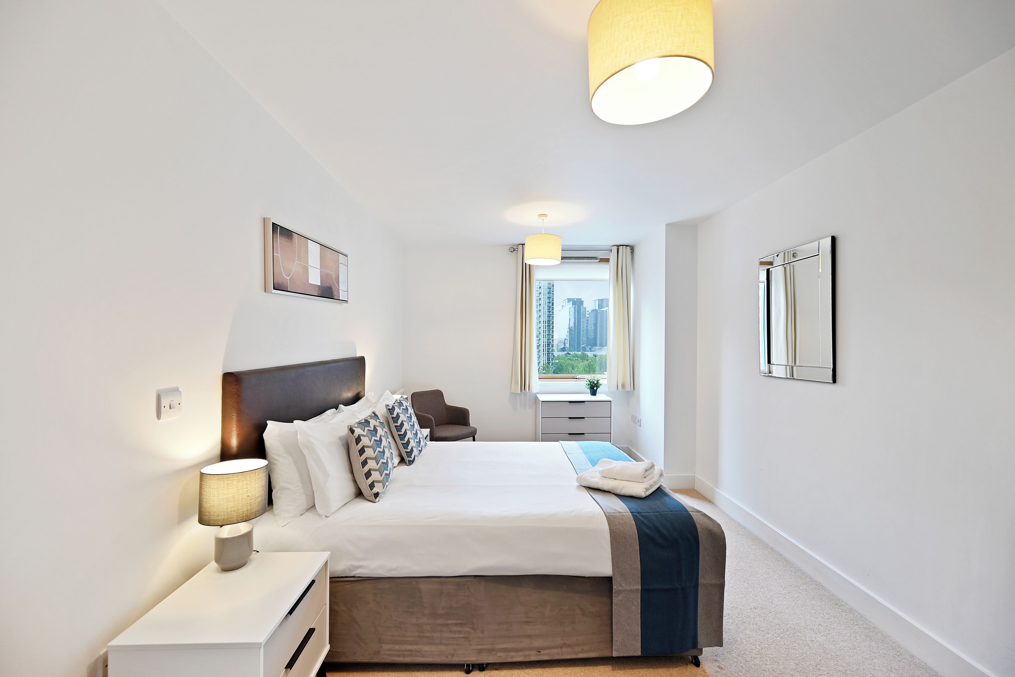 A modern hotel room with a large bed, bedside table, lamp, and a window with a city view.