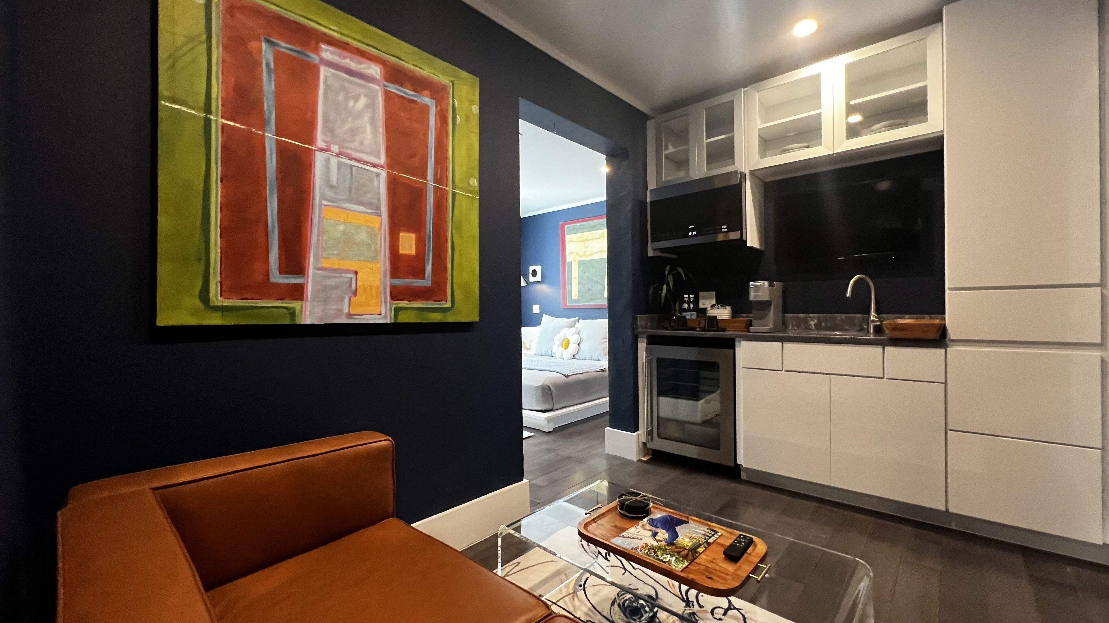 A modern kitchen with white cabinets and a built-in oven. There is a dining area with a glass table and a brown sofa. A colorful abstract painting is on the wall.