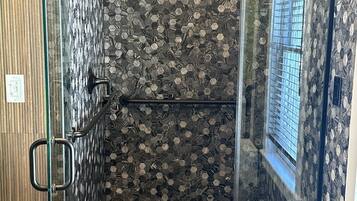 Una ducha con cerramiento de vidrio, pared con diseño de piedra y ventana a la derecha.