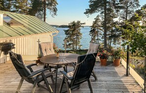 Eine Holzterrasse mit rundem Tisch und Stühlen, mit Blick auf einen See, Bäume und ein Gebäude im Hintergrund.