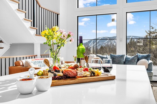 Un salon moderne doté d’une grande fenêtre, d’un escalier en bois et d’un coin repas agrémenté d’une planche de charcuterie.