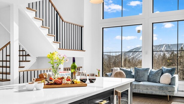 A modern kitchen with a large island, wooden staircase, and a view of mountains through large windows. - Unmatched Views of Sunday River! Slps 16! Hot Tub! (Newry)