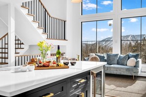 A modern kitchen with a large island, wooden staircase, and a view of mountains through large windows. - Unmatched Views of Sunday River! Slps 16! Hot Tub! (Newry)