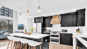 A modern kitchen with black cabinets, a white island, and a large window overlooking a forest. - Unmatched Views of Sunday River! Slps 16! Hot Tub! (Newry)