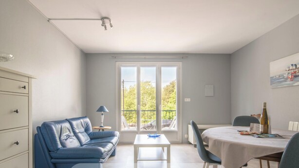 Une pièce avec un canapé bleu, une petite table, une table à manger avec des chaises, un ventilateur de plafond et une grande fenêtre donnant sur des arbres.