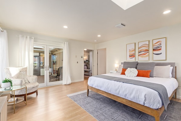 A modern bedroom with a large bed, a glass door leading to a balcony, and a small seating area with a table and chair.