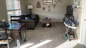 A living room with a sofa, coffee table, and TV stand. There is a window with blinds, a lamp, and wall decorations including a 'welcome' sign and a clock.