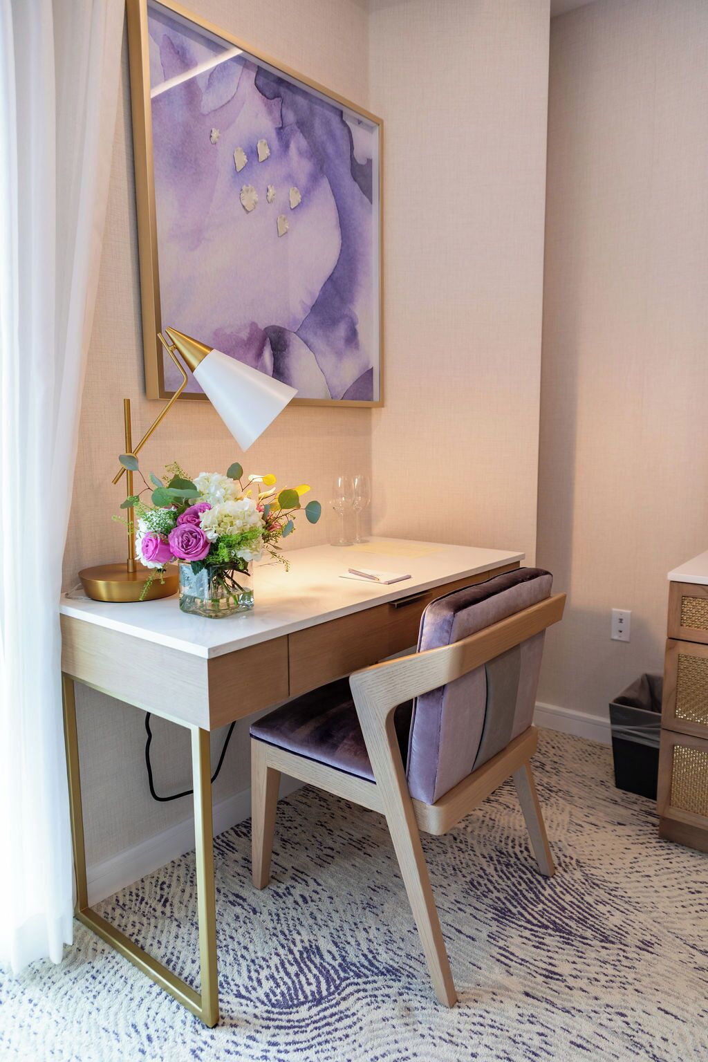 Una sala de estudio moderna con un escritorio blanco, una lámpara dorada, un jarrón con flores y un cuadro abstracto grande en la pared.