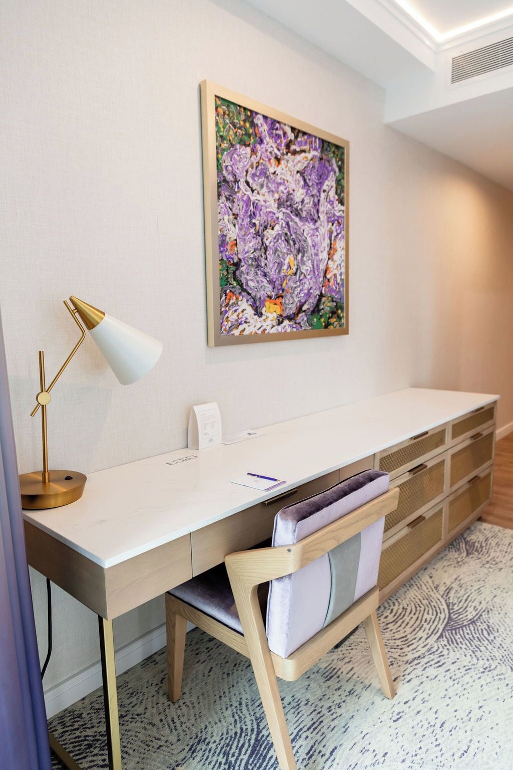A modern office space with a white desk, a gold lamp, a cushioned chair, and a colorful abstract painting on the wall.