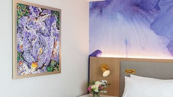 A hotel room with a bed, a nightstand with a lamp, a floral arrangement, and a large abstract painting on the wall.
