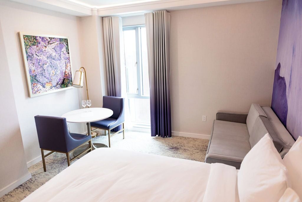 A hotel room with a bed, a round table with two chairs, a painting on the wall, and a window with curtains.