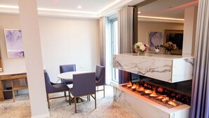 Una habitación de hotel moderna con chimenea de mármol, zona de comedor con una mesa redonda y sillas, y un ventanal con cortinas.