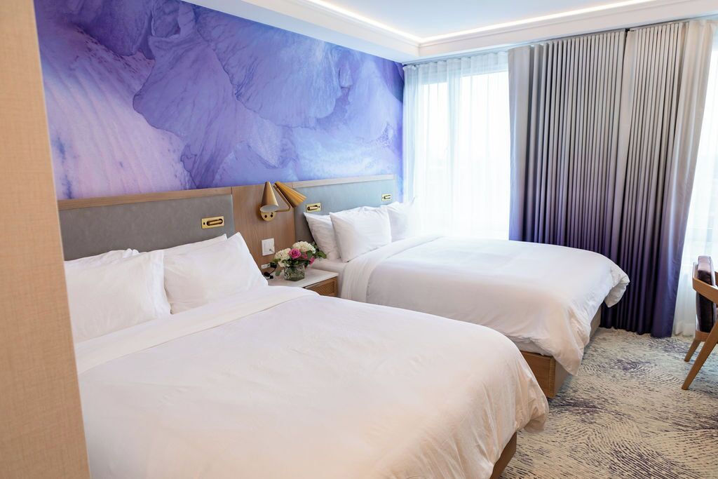 A hotel room with two beds, a headboard with a built-in shelf, a bedside table with a lamp, and a large abstract painting on the wall.