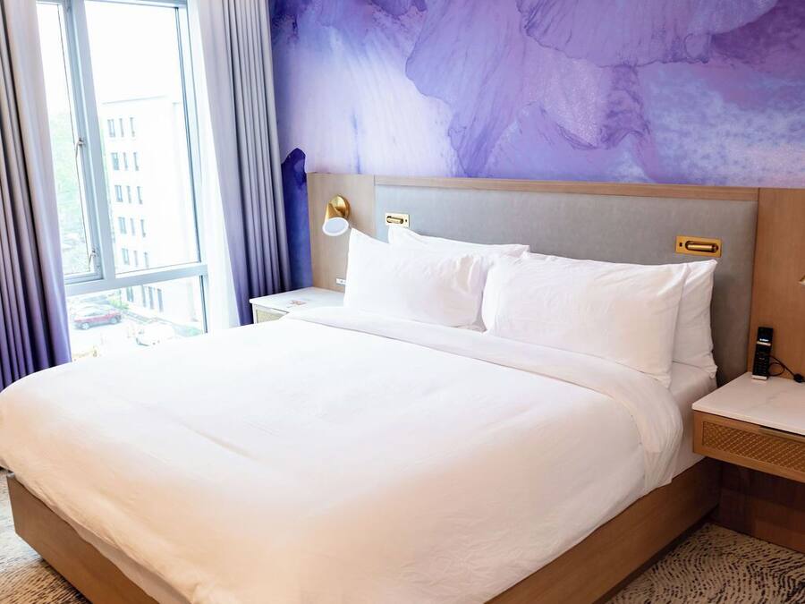Una cama grande con sábanas blancas, cabecera con acabado en madera clara y alfombra texturizada.