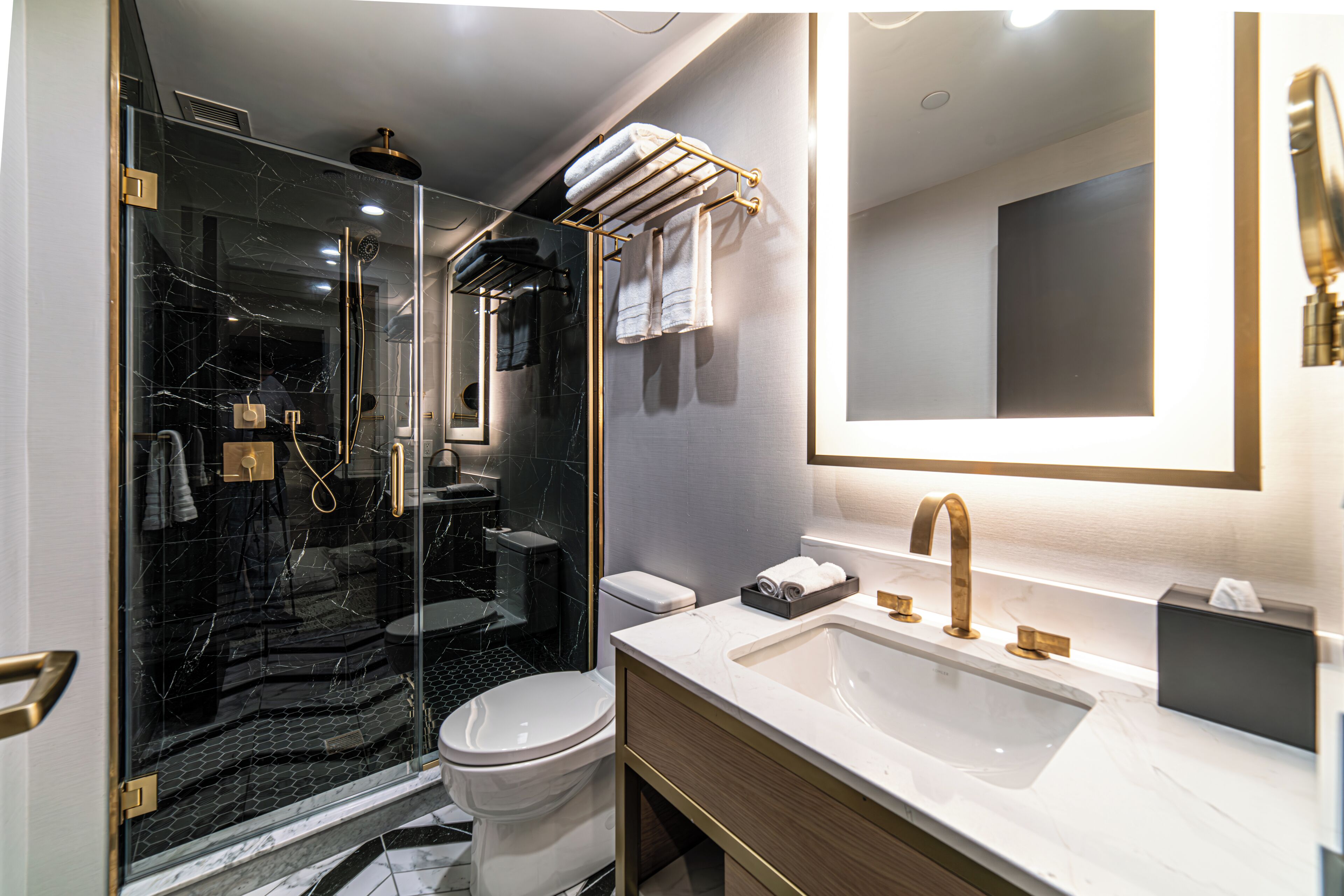 A modern bathroom with a glass-enclosed shower, a white toilet, a large sink with gold fixtures, and a mirror with integrated lighting.