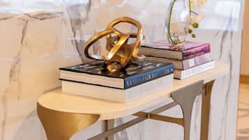 Uma parede de mármore com um espelho em moldura dourada, um objeto decorativo dourado e um vaso com flores brancas sobre uma mesinha.