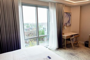A hotel room with a large window, a bed, a desk with a chair, and a painting on the wall. - Iris Hotel Boston (Brookline)