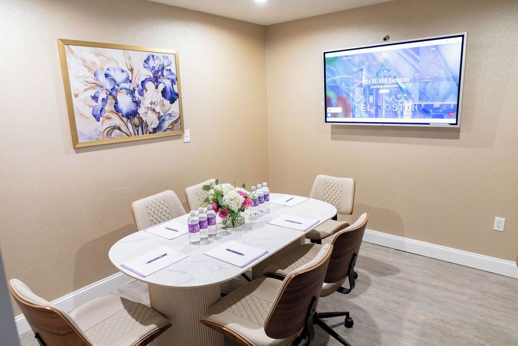 A meeting room with a long table, six chairs, a floral centerpiece, a screen displaying hotel information, and a framed painting on the wall.
