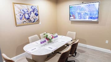 A meeting room with a long table, six chairs, a floral centerpiece, a screen displaying hotel information, and a framed painting on the wall.