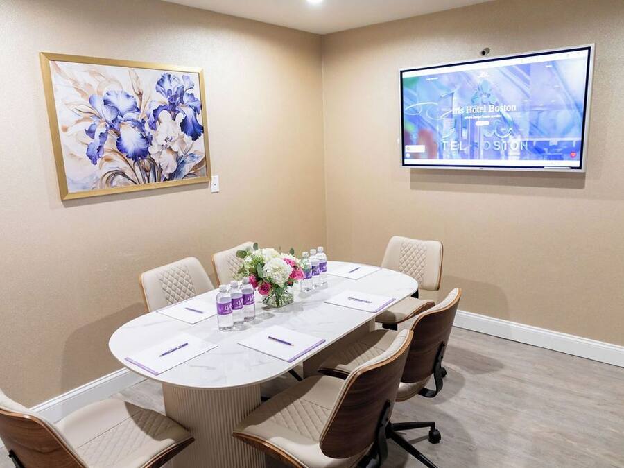 A meeting room with a long table, six chairs, a floral centerpiece, a screen displaying hotel information, and a framed painting on the wall.