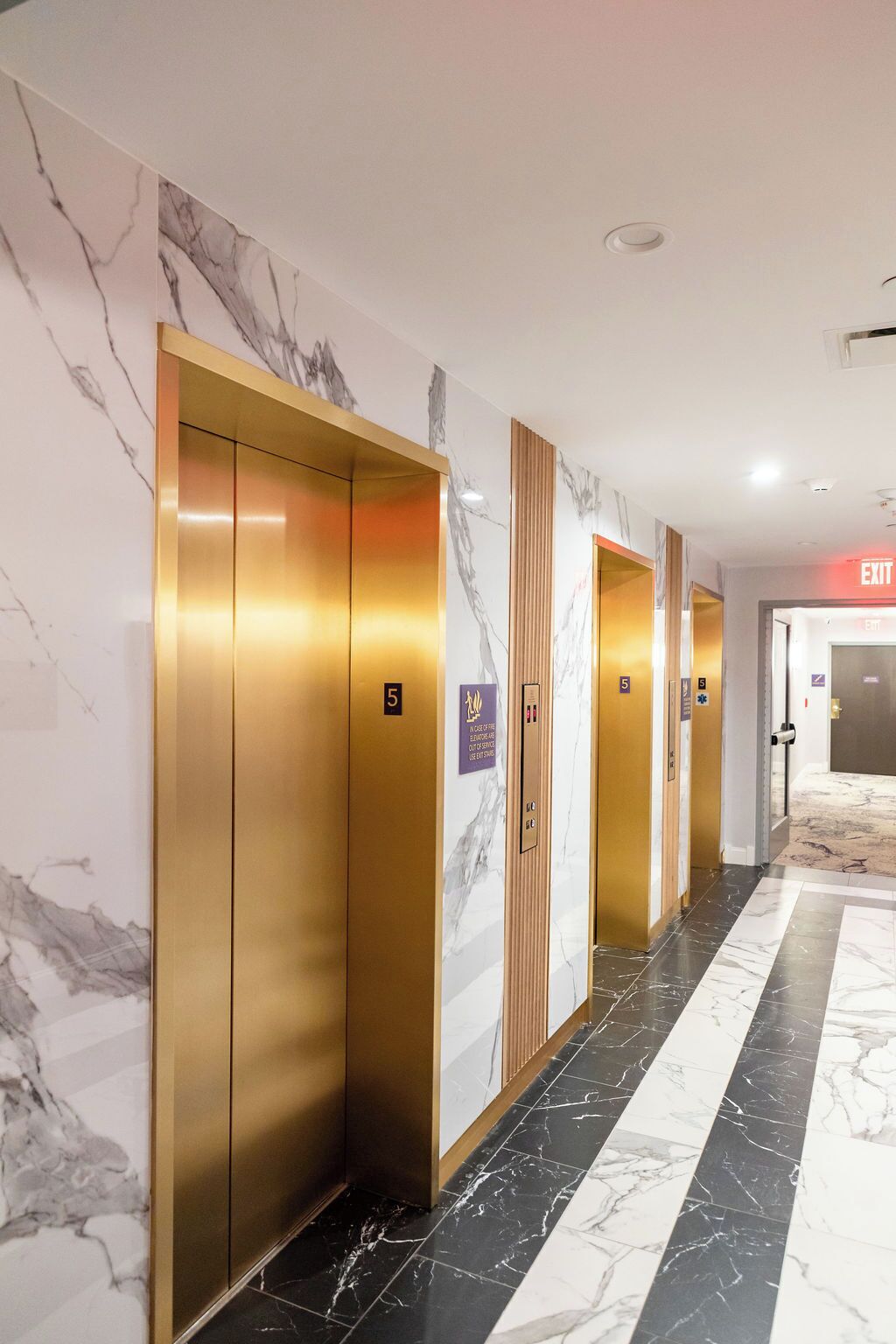 A hallway with marble walls and floors, featuring elevators and an exit sign.
