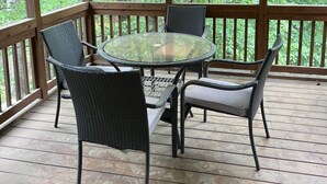 Una terraza al aire libre techada con una mesa de vidrio y sillas de mimbre, rodeada de barandas de madera y vista a un área boscosa.