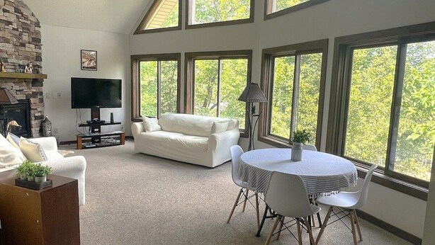 A spacious living room with a fireplace, white furniture, a TV, and large windows offering a view of trees. - Lovely Views - Top of the Mountain - Foosball & Pool Table - Screened in Porch (McGaheysville)